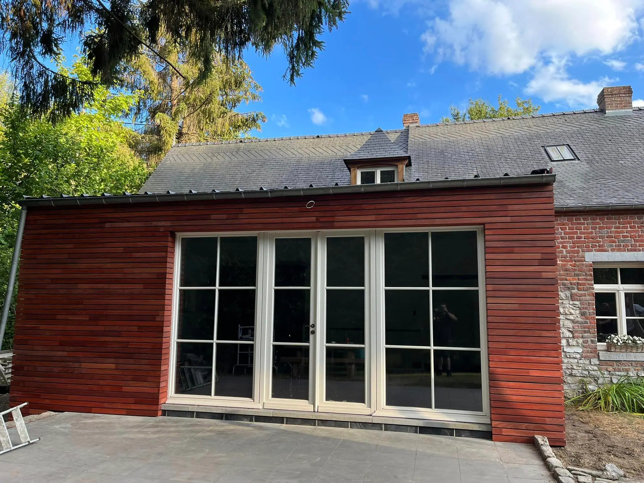 Façade entièrement bardée de clins de bois brun-rouge, avec une grande baie et un ciel bleu
