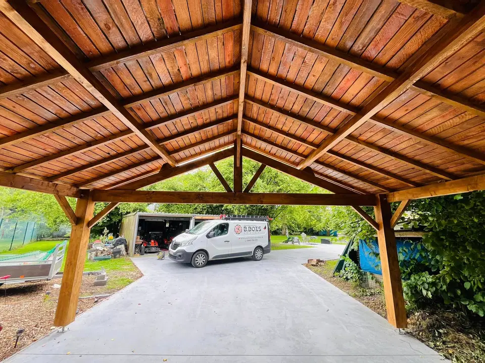 Vue par-dessous d’un carport en ferme à poinçon : charpente et sous-face en bois verni, ouverture sur l’allée
