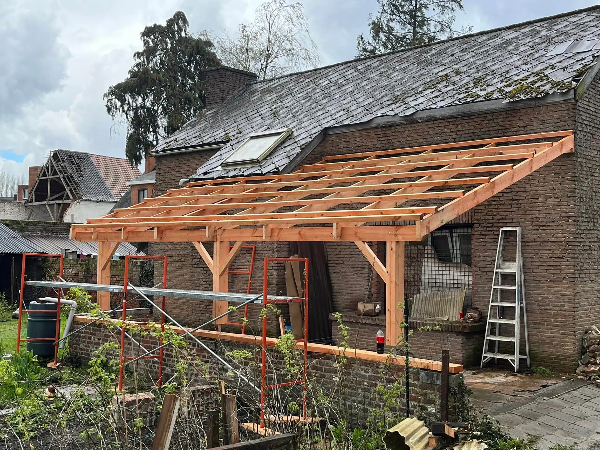 La charpente d’un carport en poteau-poutre contre un mur de brique, chevrons neufs à contre-jour