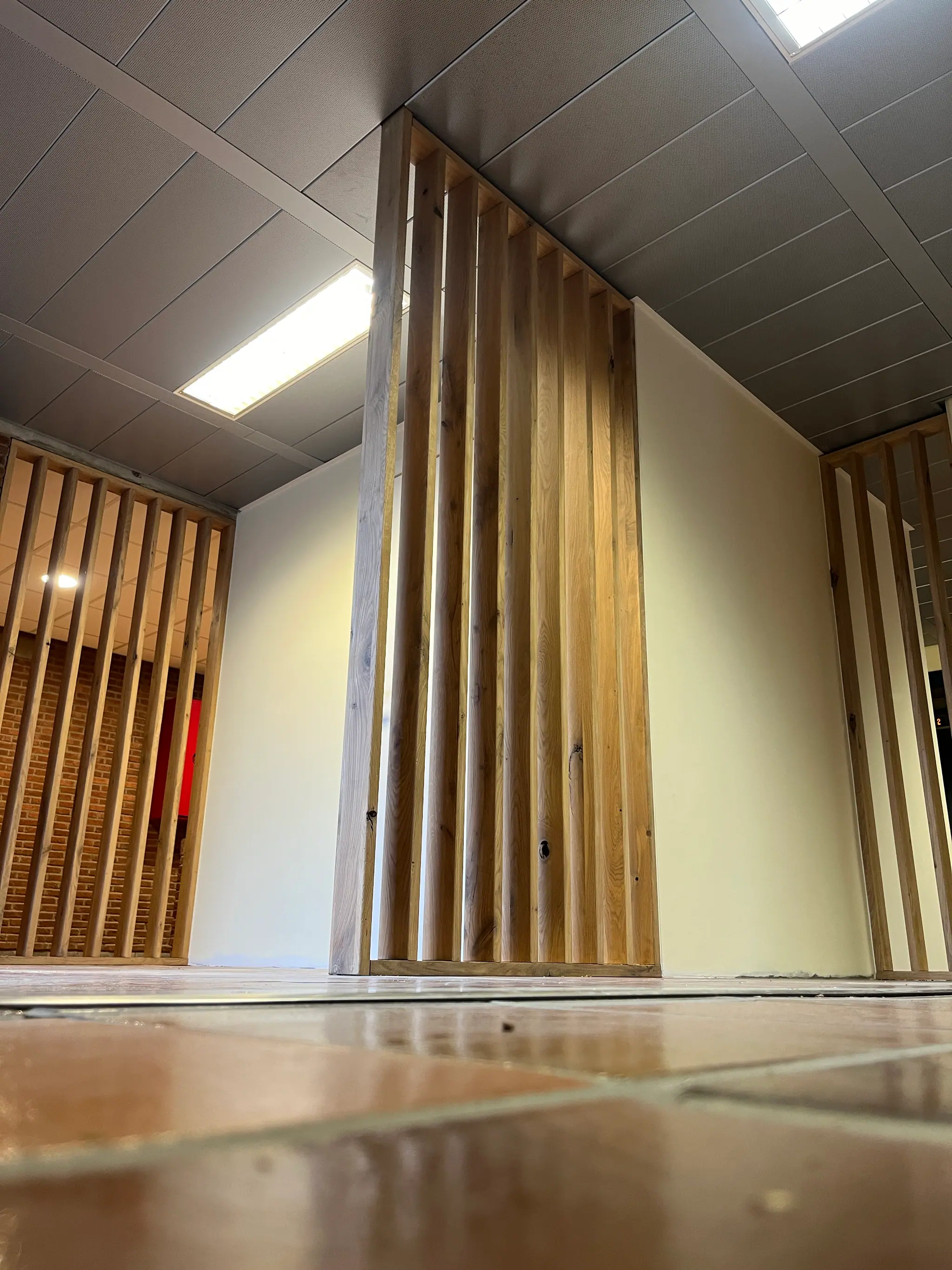 Un claustra de chêne massif traversant un couloir de bureaux, lames verticales et lumière rasante