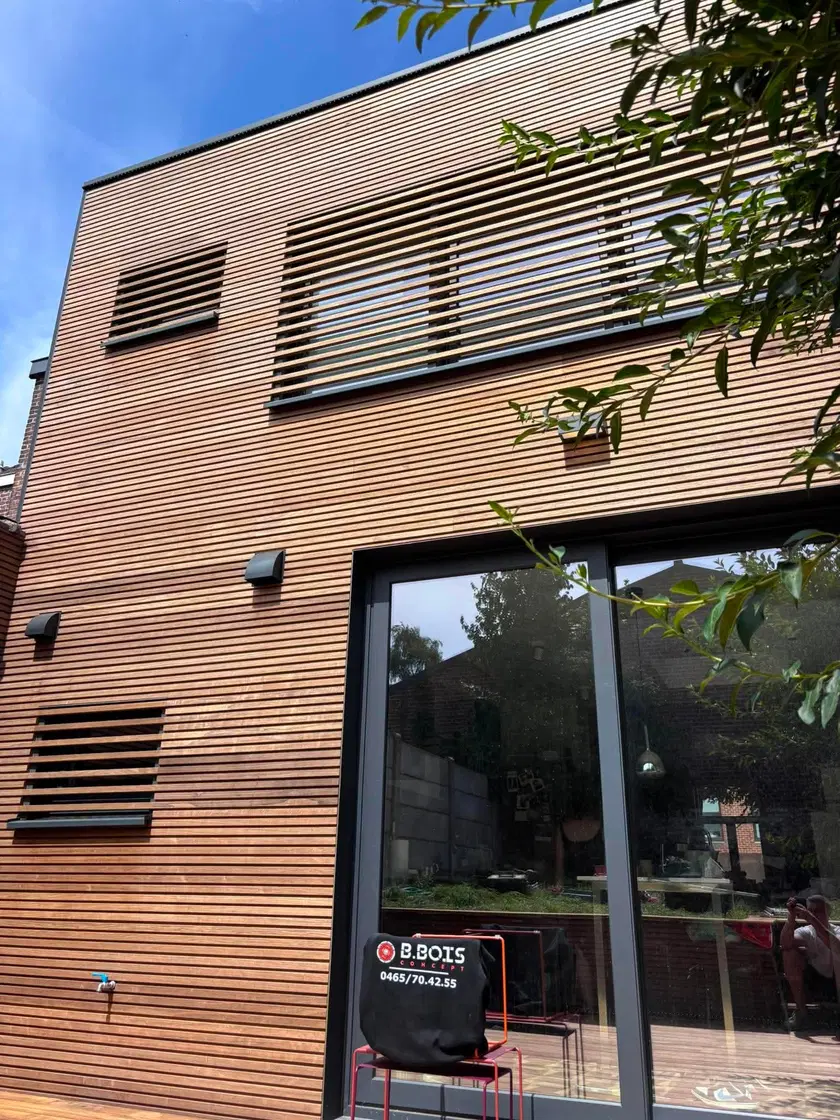 L’extension vue du jardin : bardage à lames horizontales brun-rouge, panneaux de ventilation persiennés et grandes baies à cadre noir