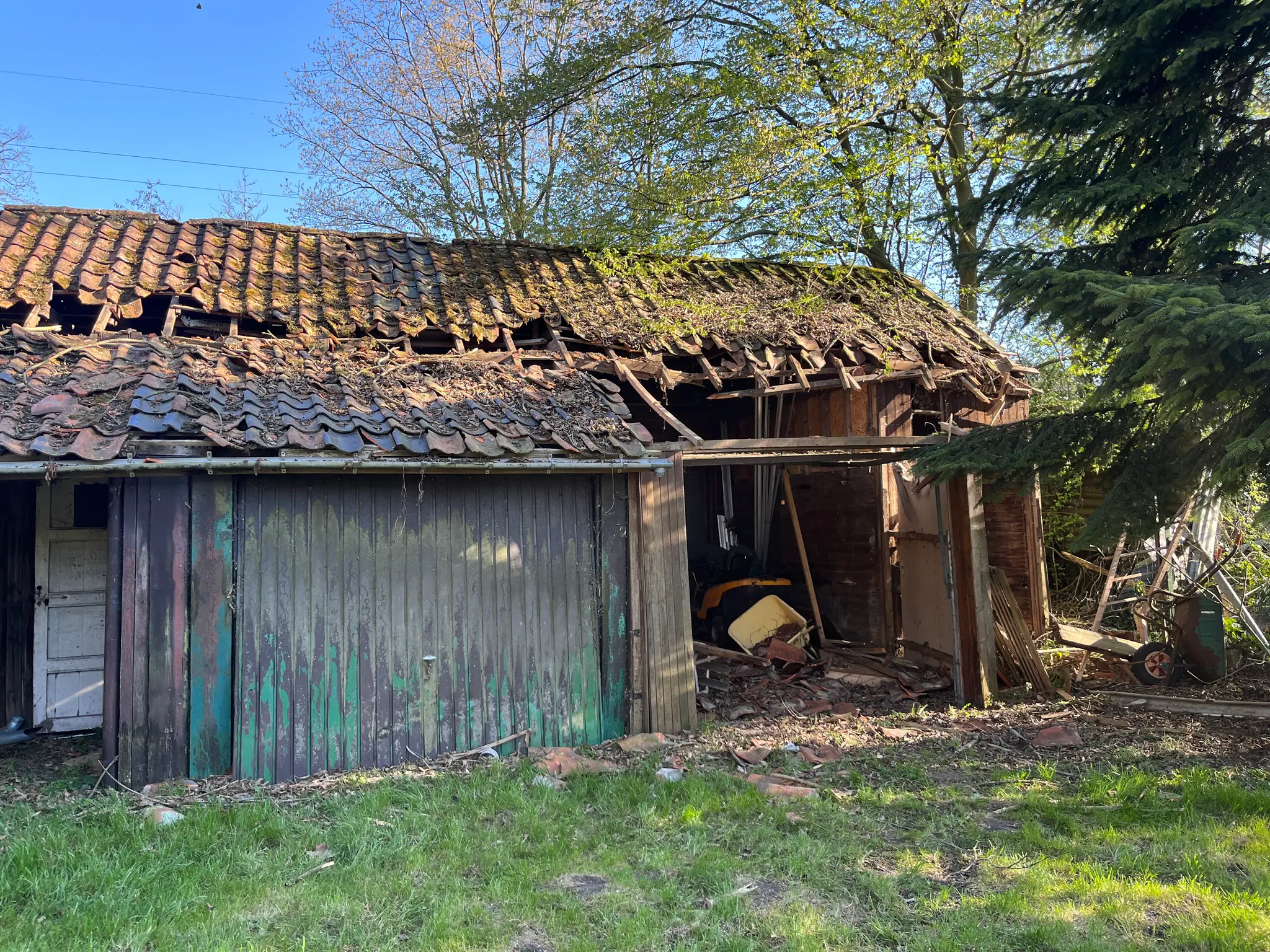 Une grange dont la toiture s’est effondrée : tuiles à moitié tombées, lattes à nu, portes de bois vert