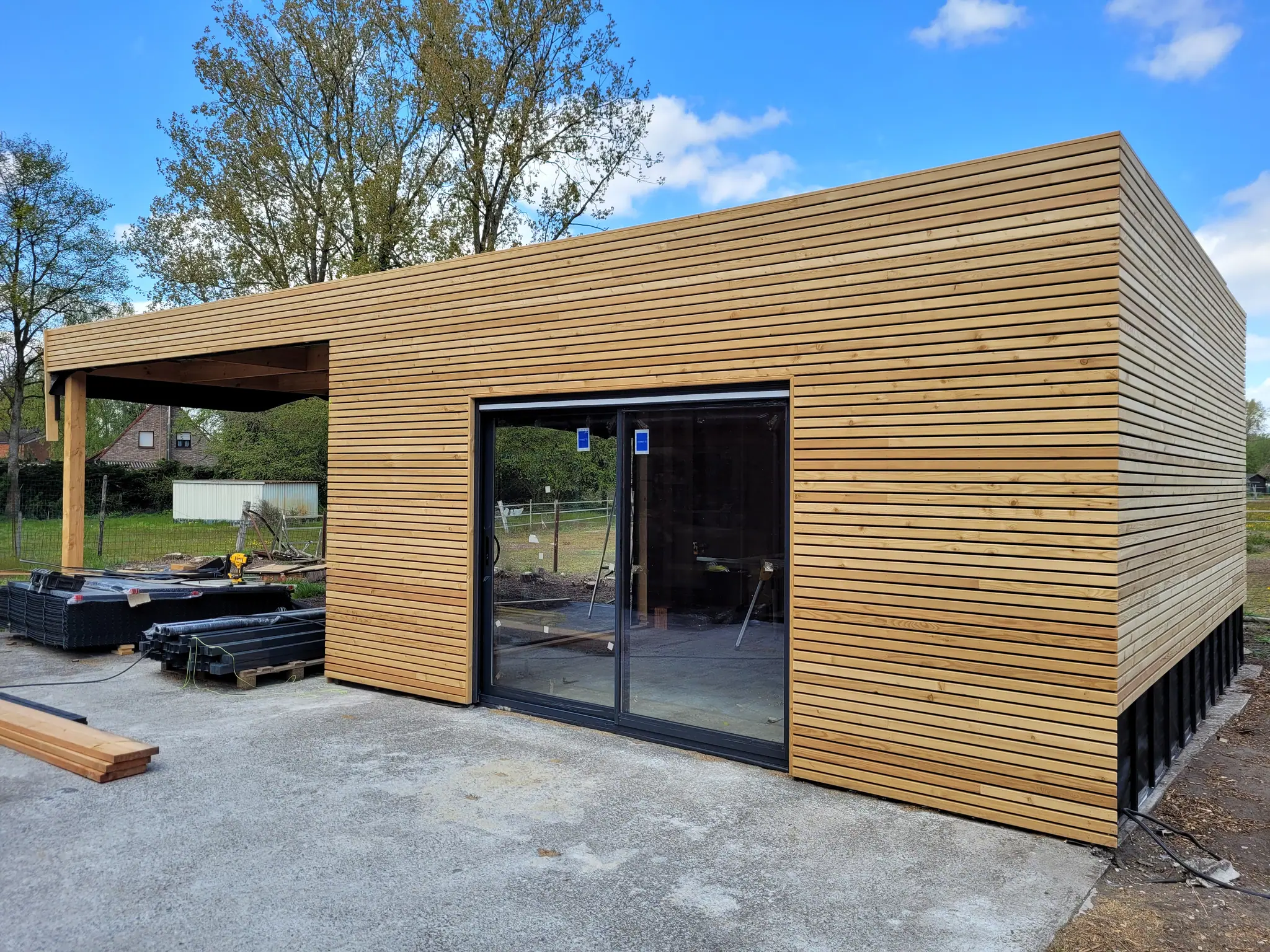 Le pavillon achevé vu de face : bardage à tasseaux horizontaux, grande baie coulissante, soubassement noir et carport ouvert sur la gauche