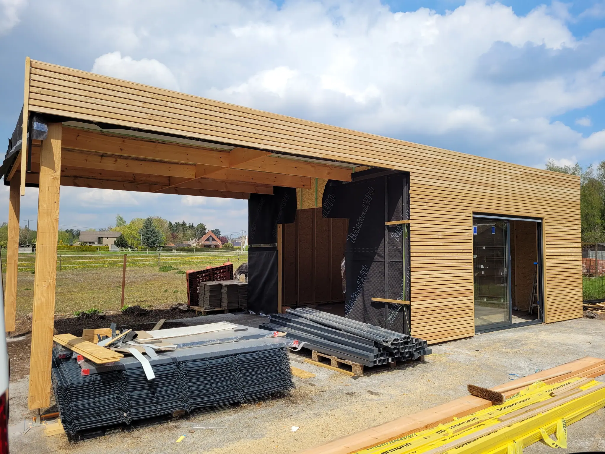 Le carport vu du dessous : poutres apparentes, sous-face habillée, et le pré au-delà