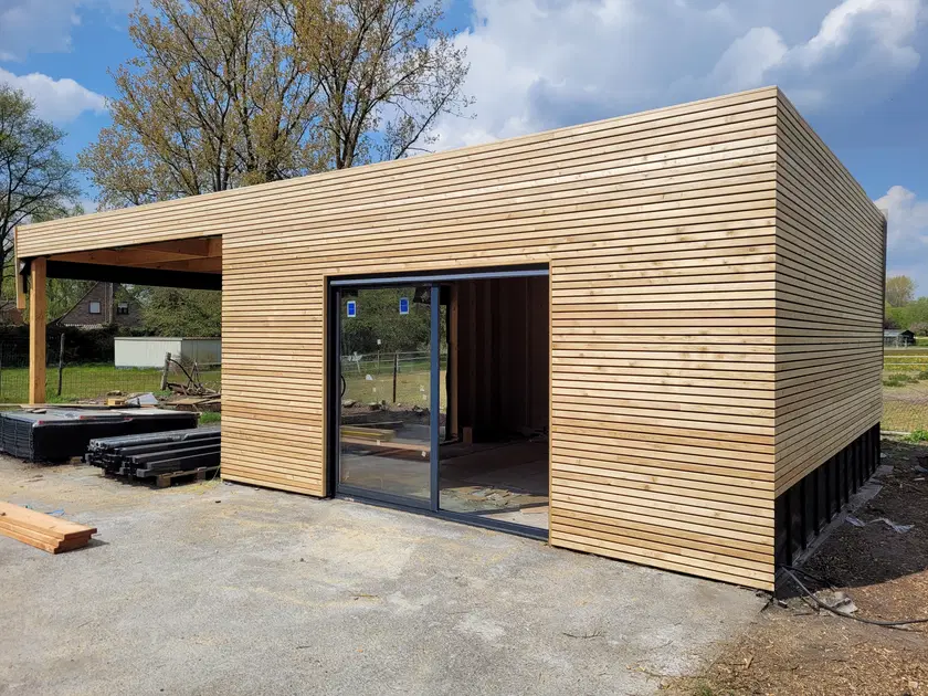 Le pavillon fini vu de trois quarts : bardage à tasseaux horizontaux, toiture plate, carport ouvert sur la gauche et grande baie coulissante