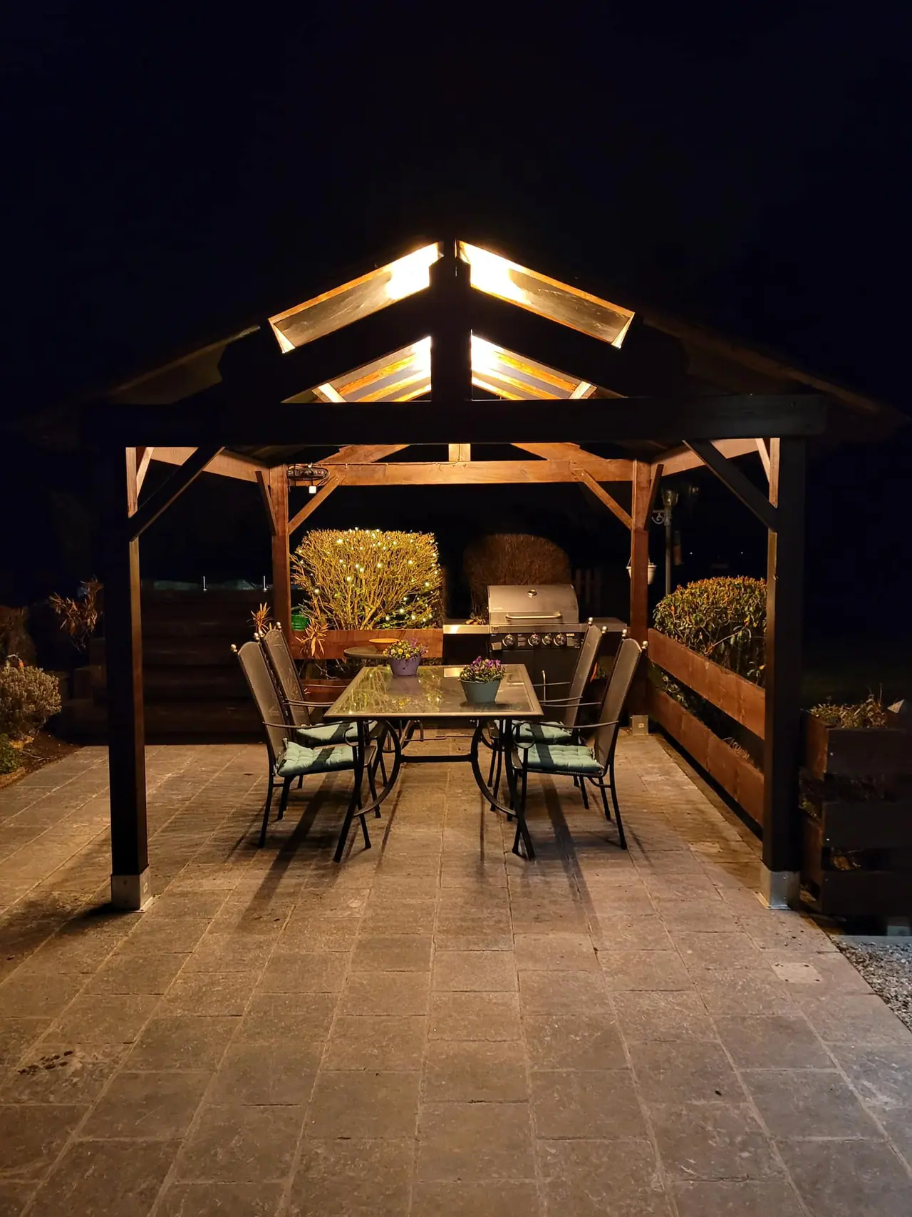 Pergola en bois éclairée le soir, ferme à poinçon apparente au-dessus d’une table dressée