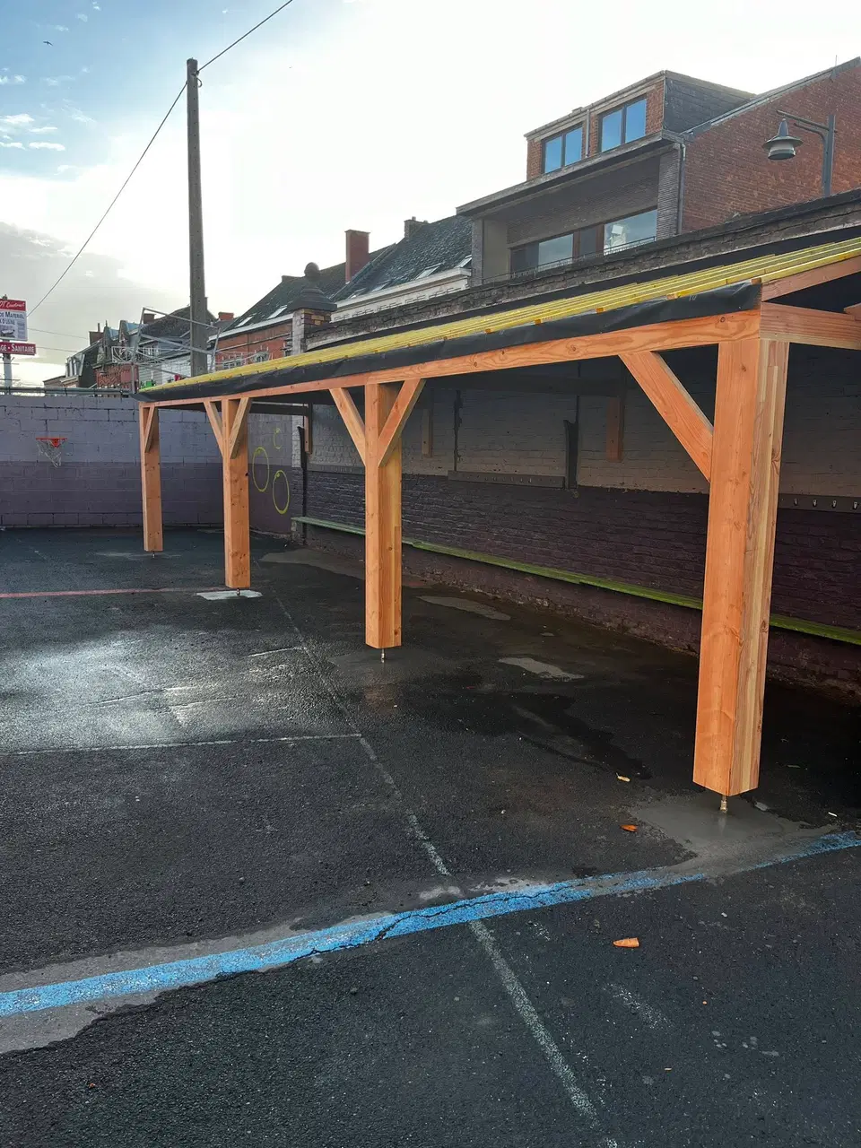 Préau scolaire terminé vu en enfilade : charpente en douglas, couverture posée et marquages au sol de la cour