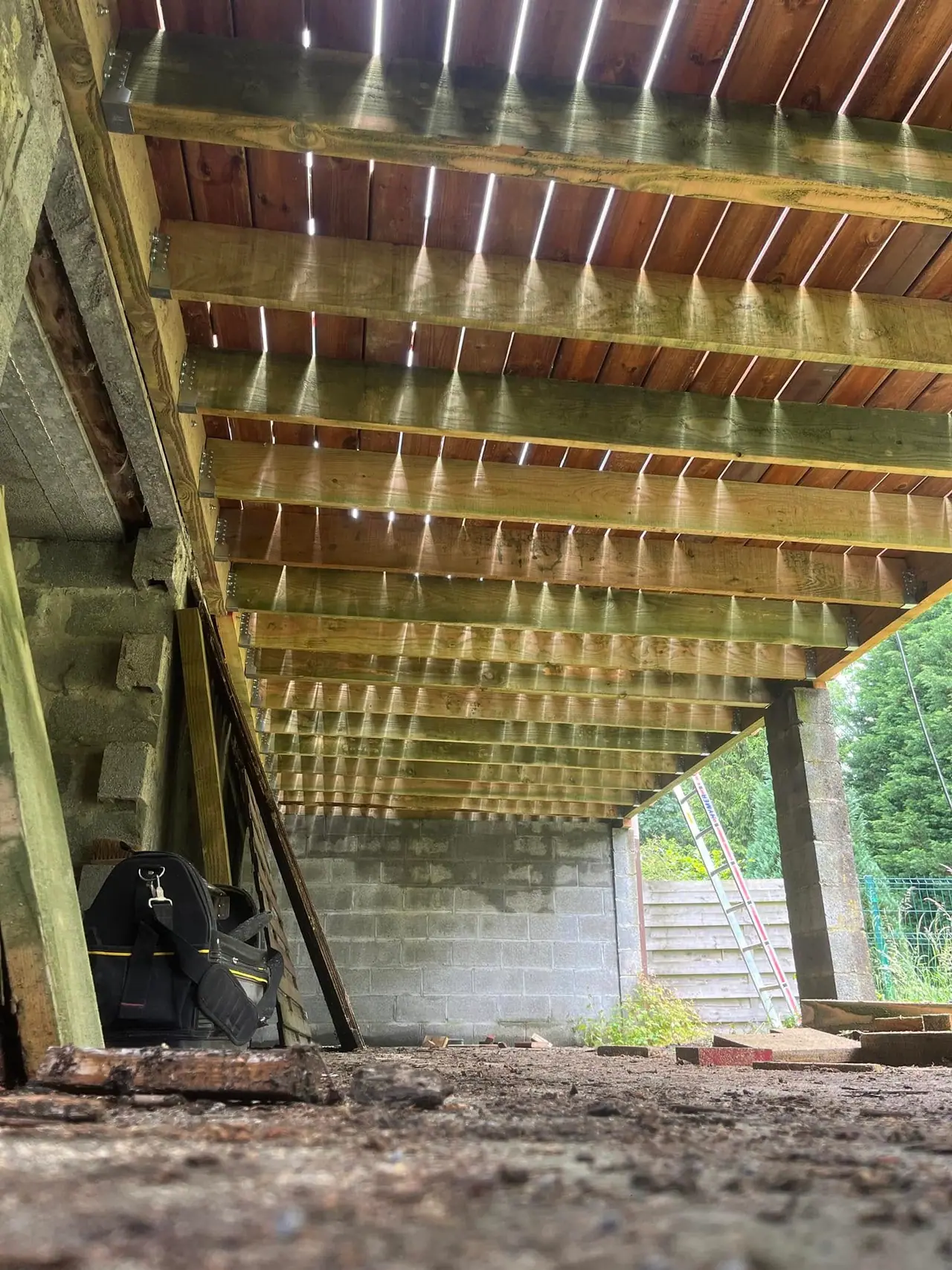 Vue de dessous d’une terrasse : structure de solives et de poteaux, lumière traversant les lames